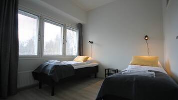 A room with two beds, a window with blinds, a small table, and a wall-mounted lamp.