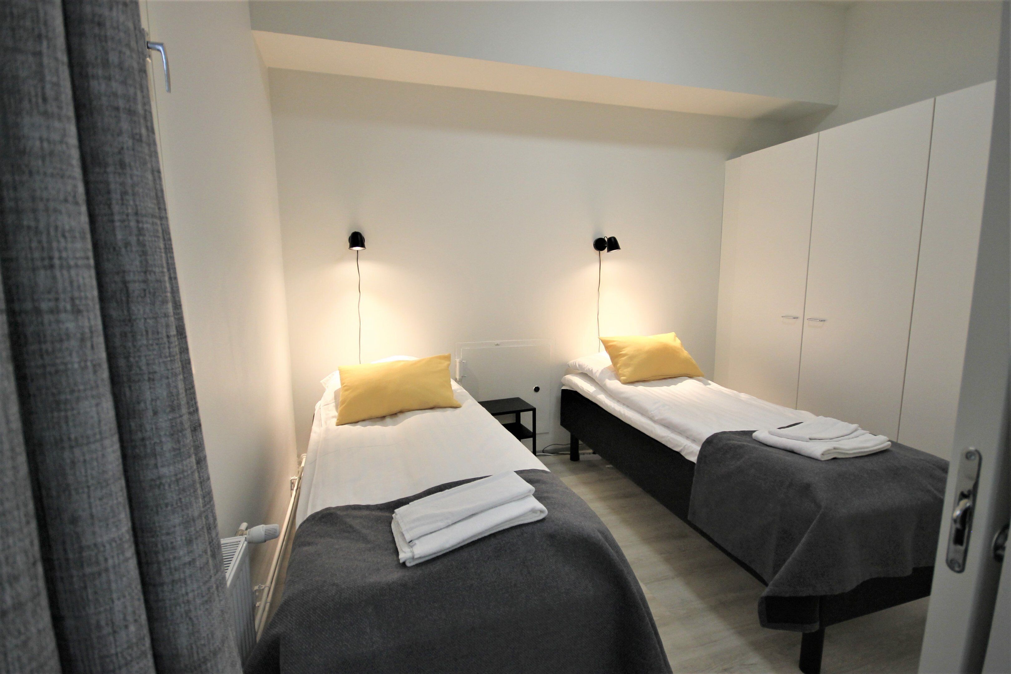 Een hotelkamer met twee eenpersoonsbedden, elk met een kussen en dekbed. Tussen de bedden staat een klein tafeltje, en er is een kledingkast op de achtergrond te zien.