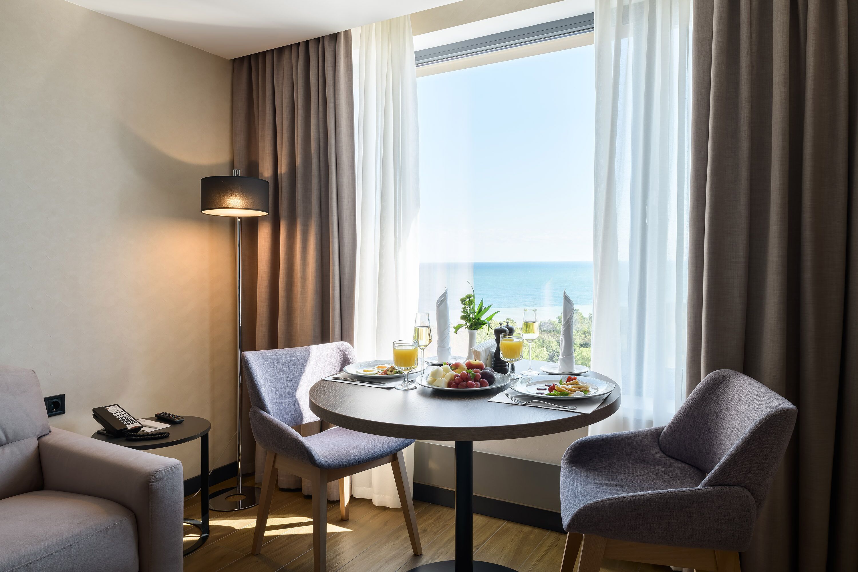 Une chambre d’hôtel avec une table à manger ronde, deux chaises, une lampe, un téléphone et une vue sur la mer.