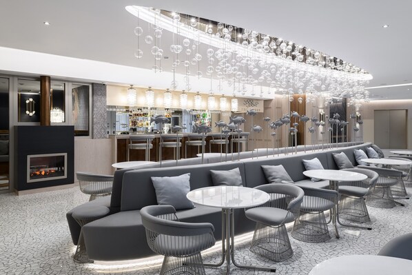 Een moderne bar met een lange toonbank, zitplaatsen met ronde tafels en stoelen, en een decoratief plafond met hangende lampen.