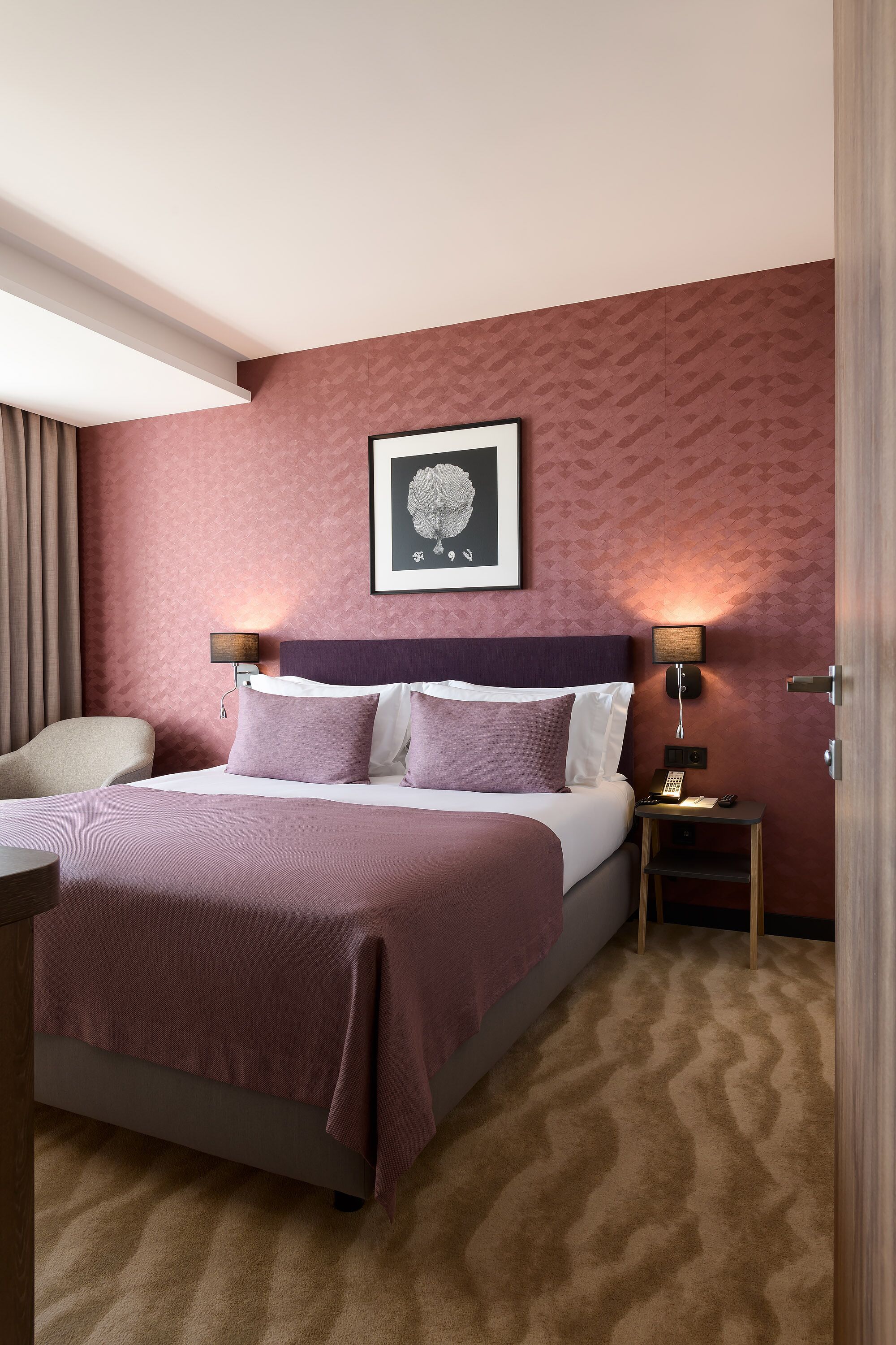 A hotel room with a large bed, two bedside tables, a chair, a framed picture on the wall, and a patterned carpet.