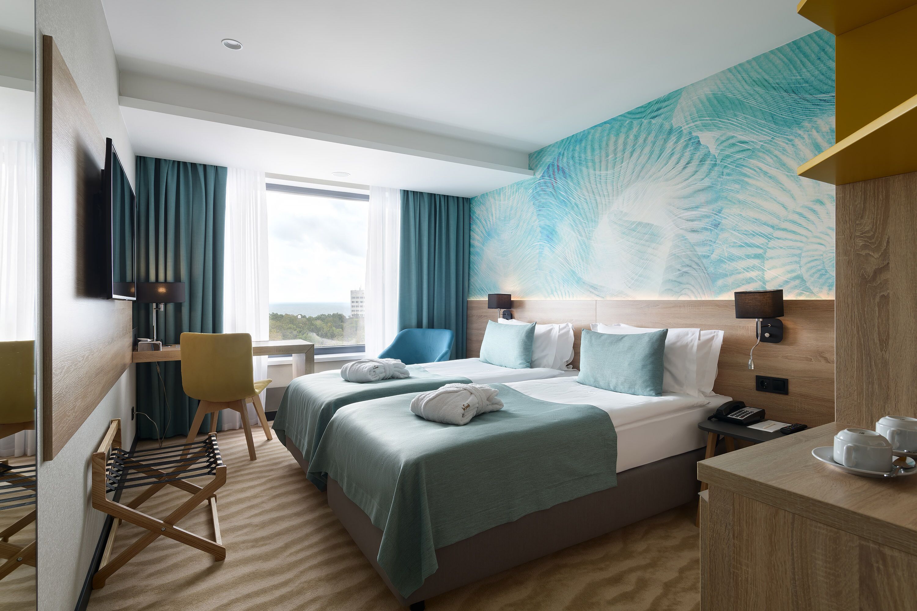 Ein Hotelzimmer mit zwei Betten, einem Schreibtisch, einem Stuhl, einem Fernseher und einem großen Fenster mit Vorhängen.