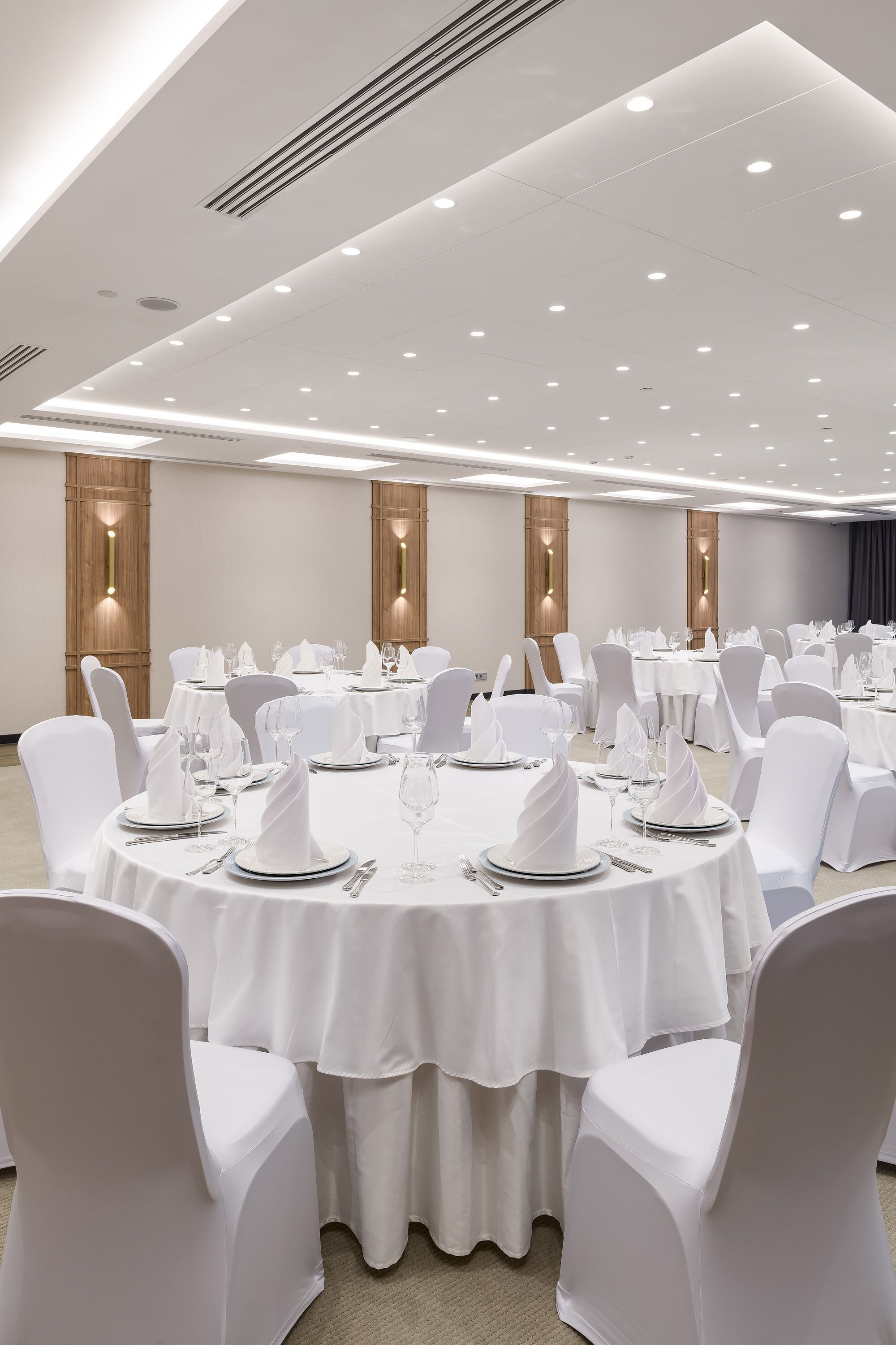 Un salón de banquetes con mesas redondas dispuestas para comer, sillas blancas e iluminación colgante.