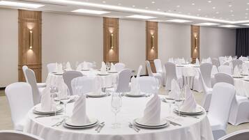 Un salón de banquetes con mesas redondas dispuestas para comer, sillas blancas e iluminación colgante.