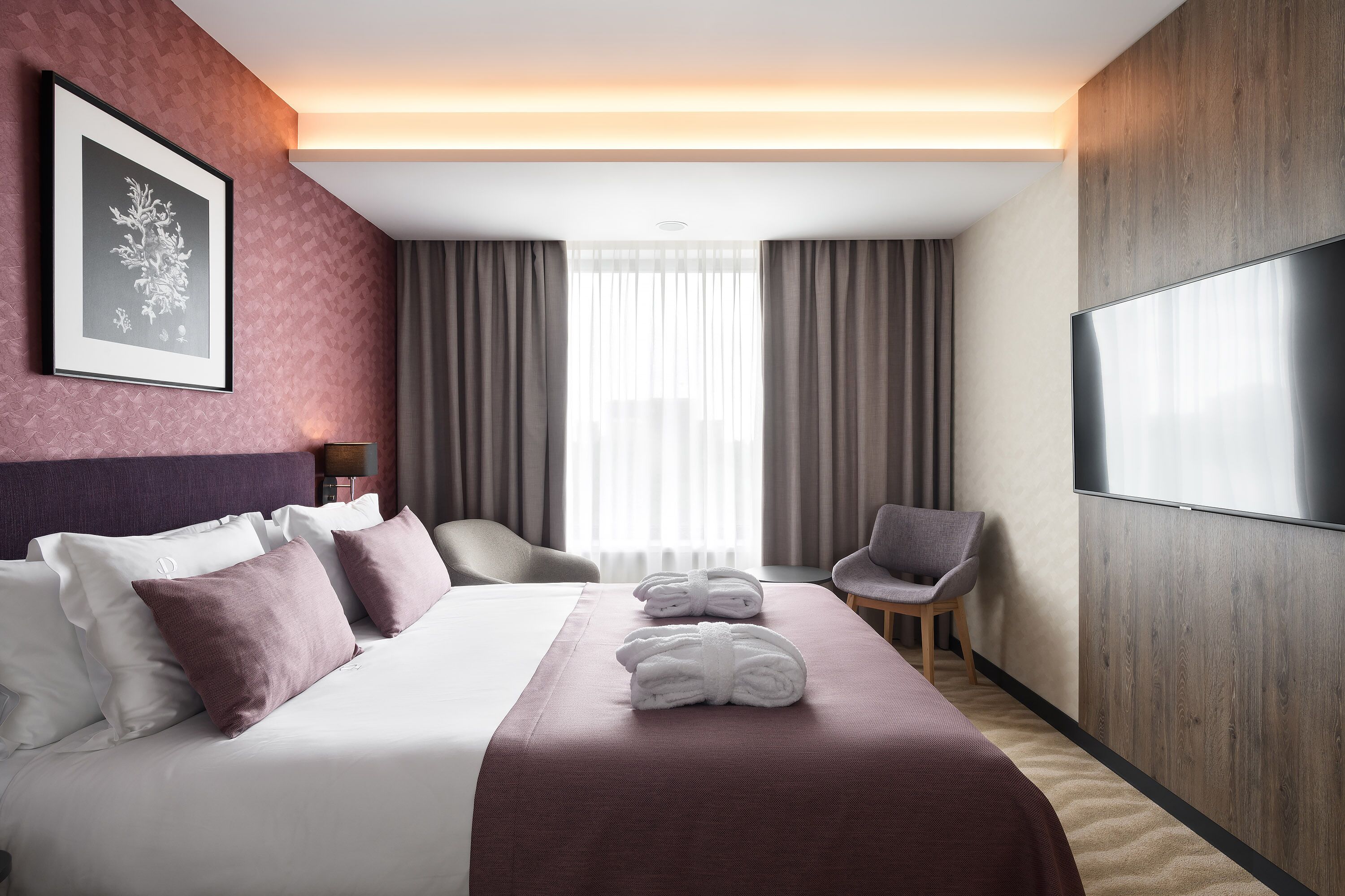 A hotel room with a large bed, a flat-screen TV, a framed picture on the wall, and a view of the city through the window.