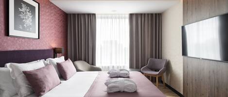 A hotel room with a large bed, a flat-screen TV, a framed picture on the wall, and a view of the city through the window.