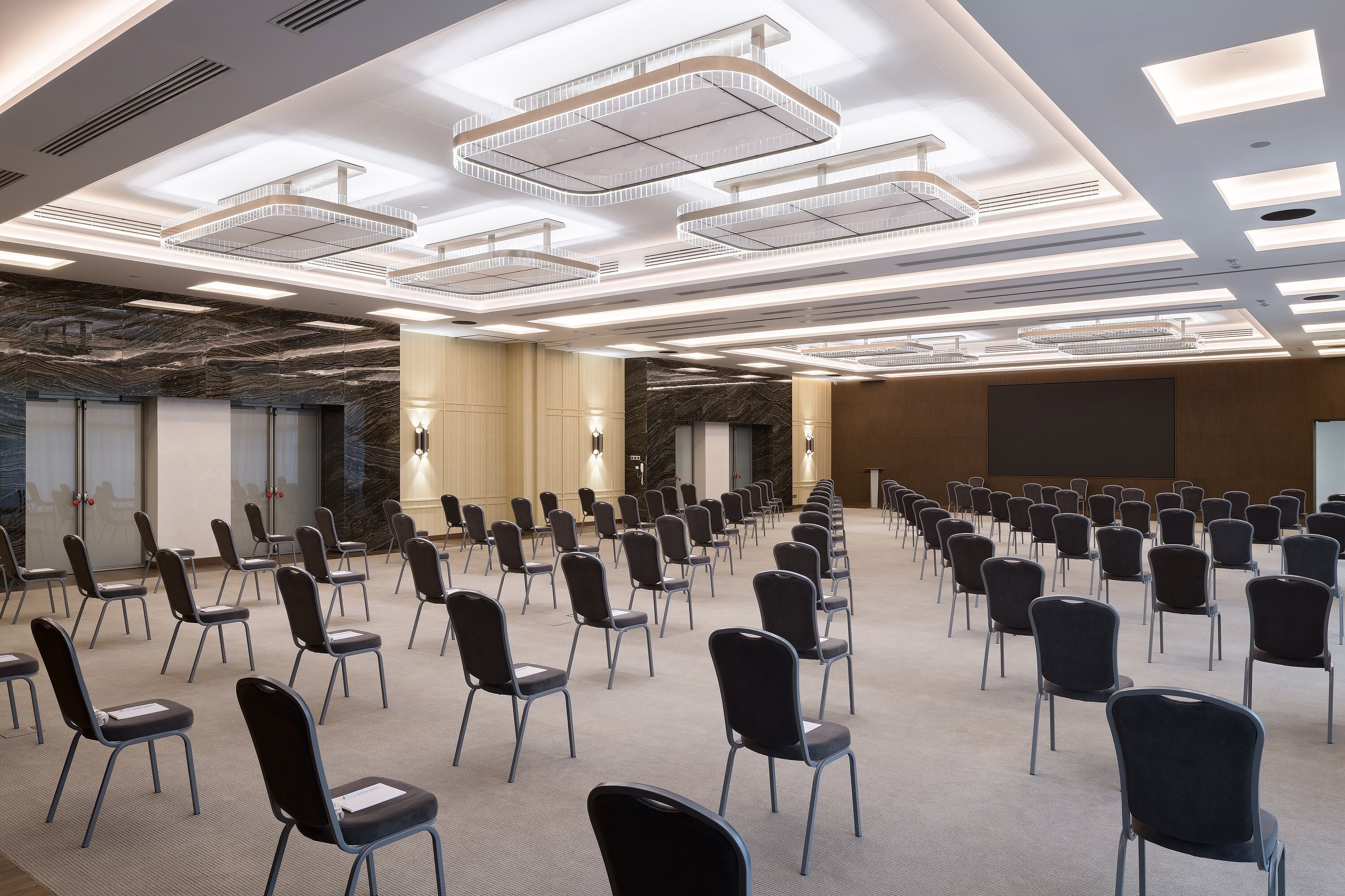 A conference room with rows of chairs, a marble wall, and a large screen.