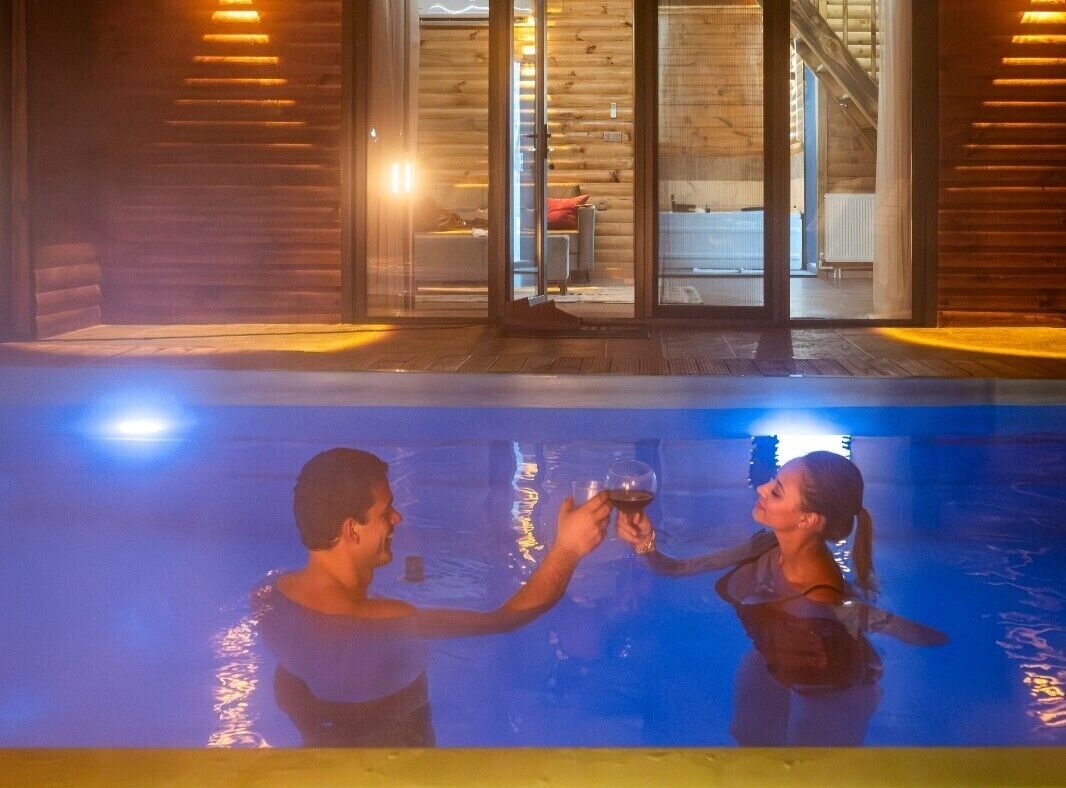 Un couple trinque au bord d’une piscine, avec une cabane en bois en arrière-plan.