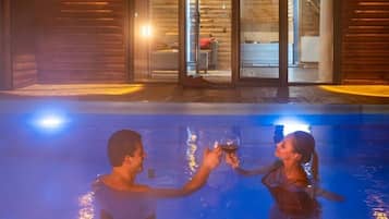 Un couple trinque au bord d’une piscine, avec une cabane en bois en arrière-plan.