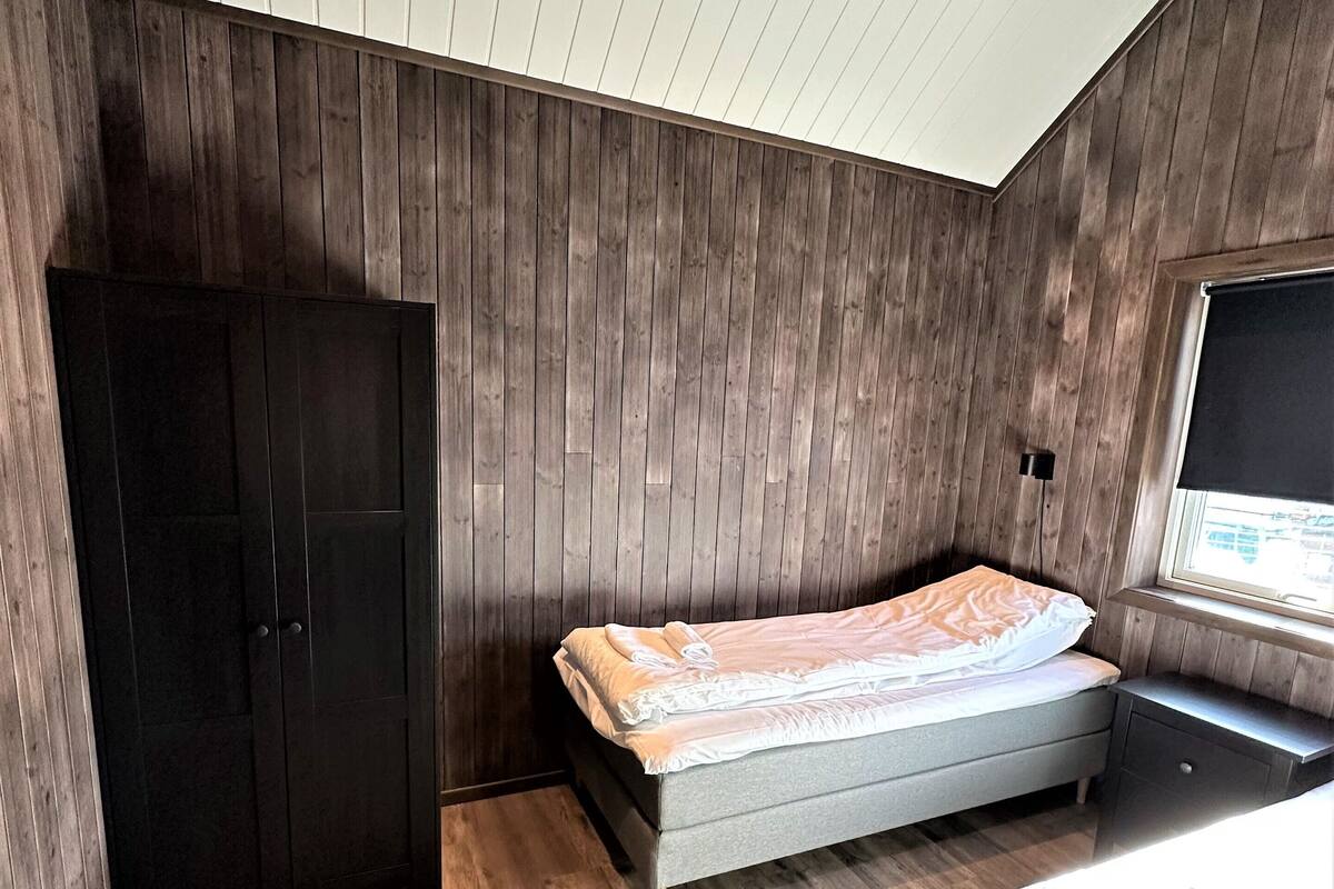 Quarto com teto inclinado, paredes de madeira, uma cama, um guarda-roupa e uma janela.