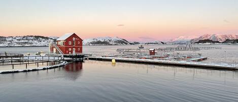 Un paysage paisible au bord de l’eau, avec un bâtiment rouge, des bateaux amarrés et, au loin, des montagnes enneigées.