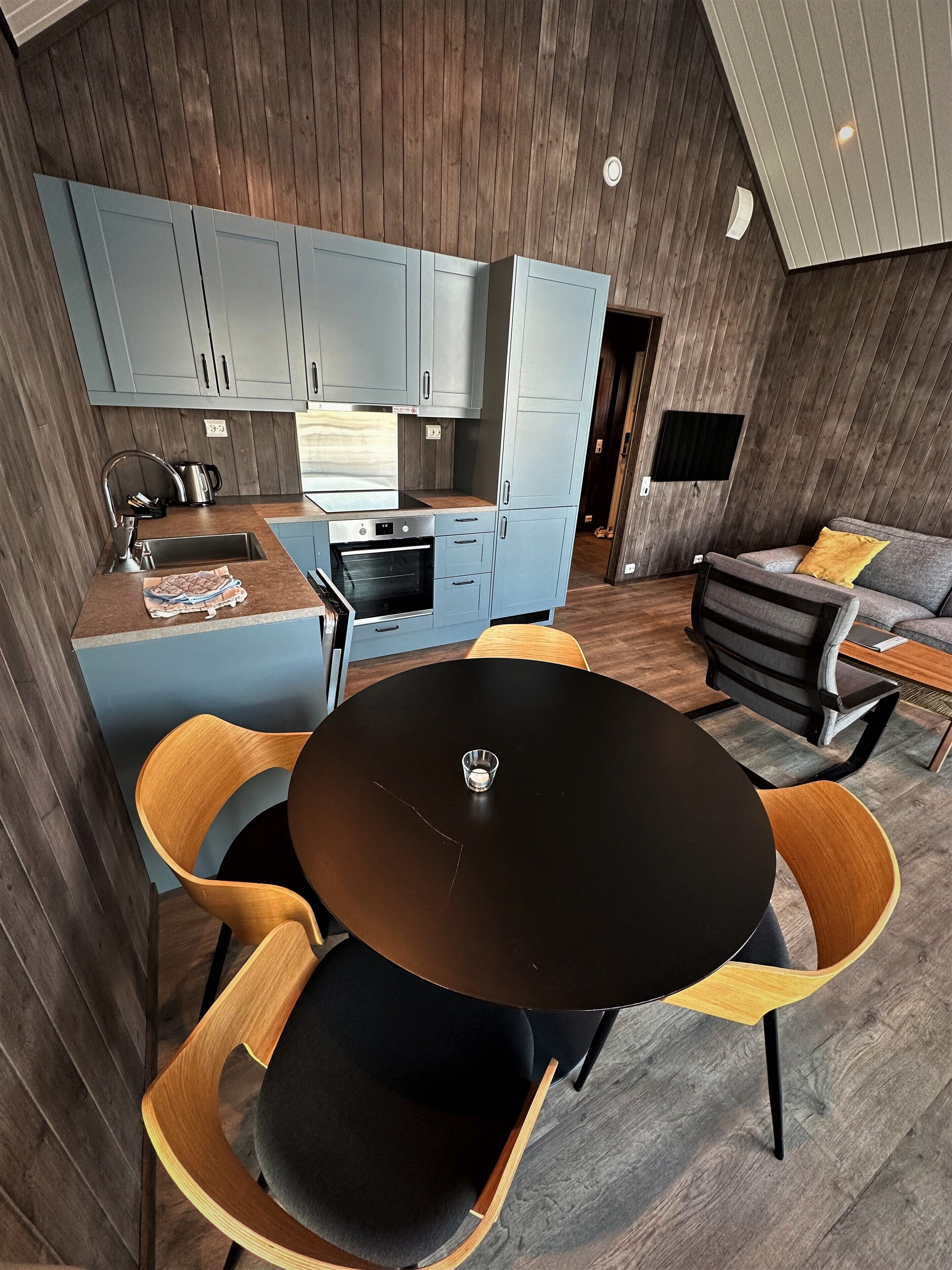 A modern kitchen with grey cabinets, a black countertop, and a built-in oven. There is a dining area with a black table and wooden chairs.