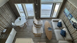 Habitación con techos altos, interior de madera, zona de comedor, sofá con cojines y vista al mar a través de amplias ventanas.