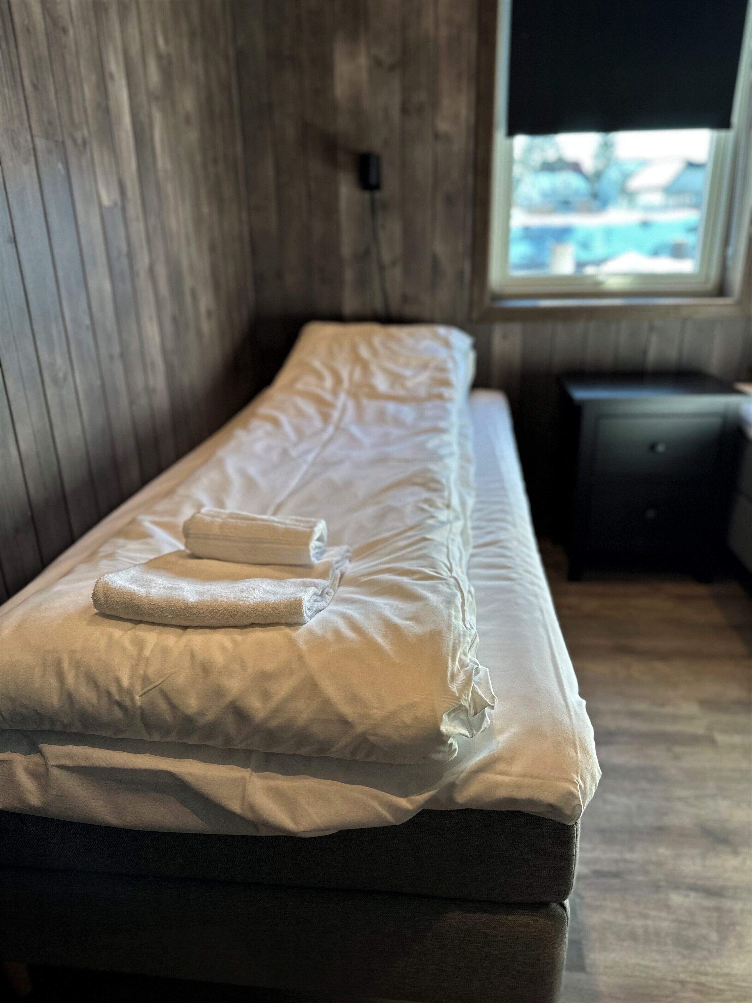 A single bed with white linens and two folded towels, a wooden wall, a small bedside table, and a window with a view.