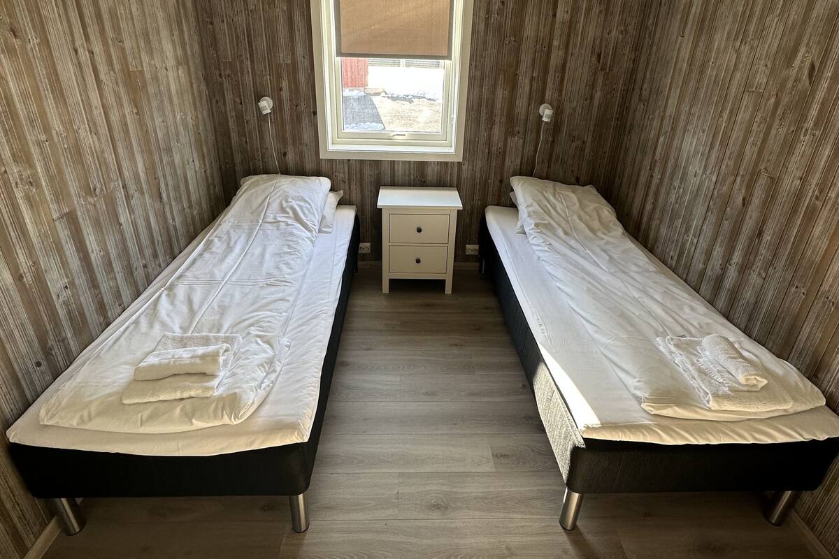 Quarto com duas camas de solteiro, uma pequena mesa de cabeceira com gavetas, uma janela com cortina e paredes revestidas de madeira.