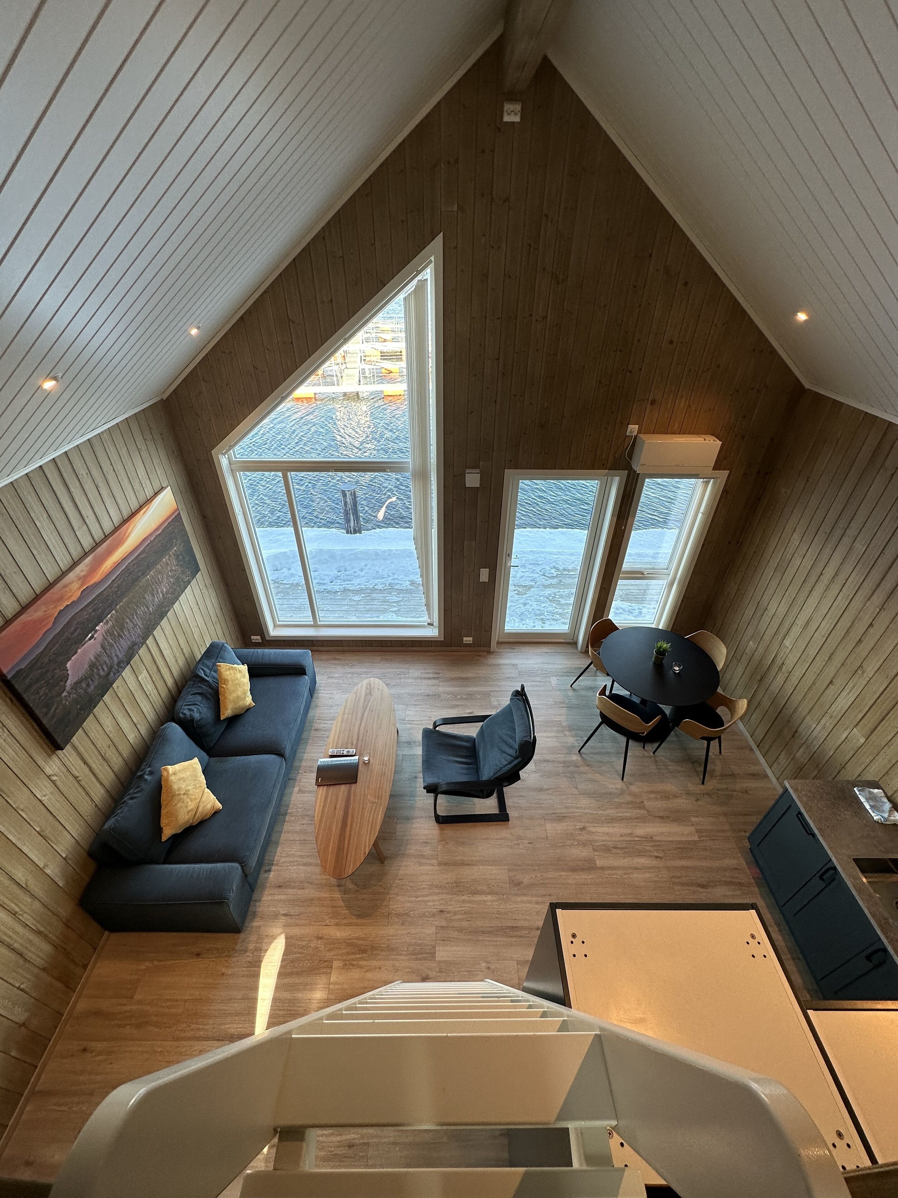 A modern loft-style room with a sloped ceiling, large windows, a sofa, a coffee table, a chair, and a dining table.