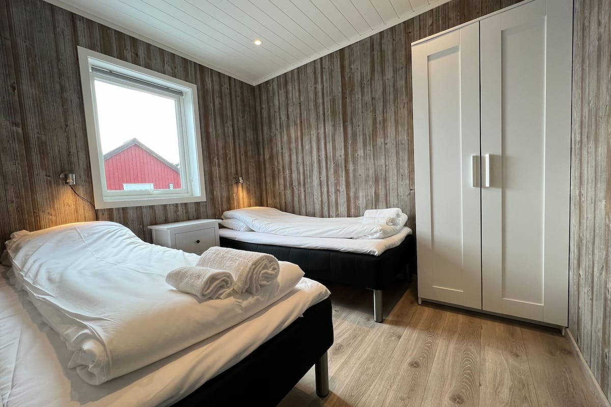 Quarto com duas camas, guarda-roupa, janela com vista para um edifício vermelho e painéis de madeira nas paredes.