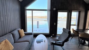 Un interior moderno con un sofá gris, una mesita redonda y una zona de comedor con mesa y sillas. Amplios ventanales ofrecen vistas al exterior.