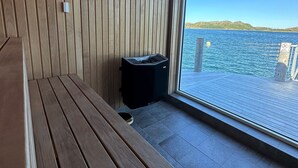 Un banco de madera y un calentador negro en una habitación con un ventanal grande que da a un cuerpo de agua.