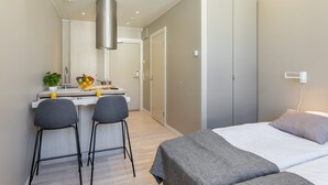 Une chambre d’hôtel moderne équipée d’un lit, d’une table de chevet et d’un petit coin cuisine avec un panier de fruits et des tabourets de bar.