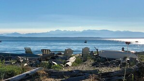 Une plage parsemée de chaises et de troncs d’arbres, un bateau sur l’eau et des montagnes en arrière-plan.