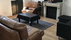 Uma sala de estar com sofá de couro, mesa de centro preta, poltrona de couro marrom, televisão de tela plana, lareira e uma grande janela.