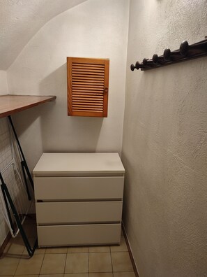 Ein kleiner, kompakter Raum mit einem Holzschrank, einer weißen Kommode, einem Wandregal mit Haken und einem Metallgestell.