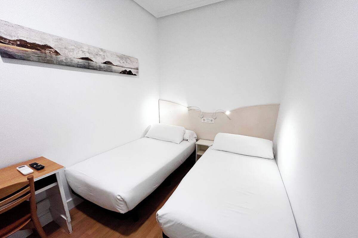 Una habitación pequeña y limpia con dos camas individuales, un escritorio y una mesita de noche.