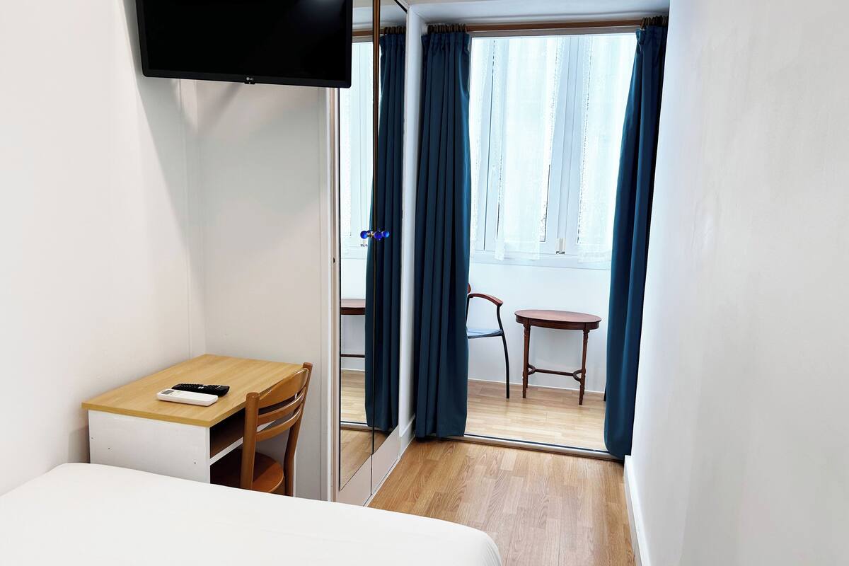 Una habitación de hotel con una cama, un escritorio con silla, un televisor montado en la pared y una ventana con cortinas.