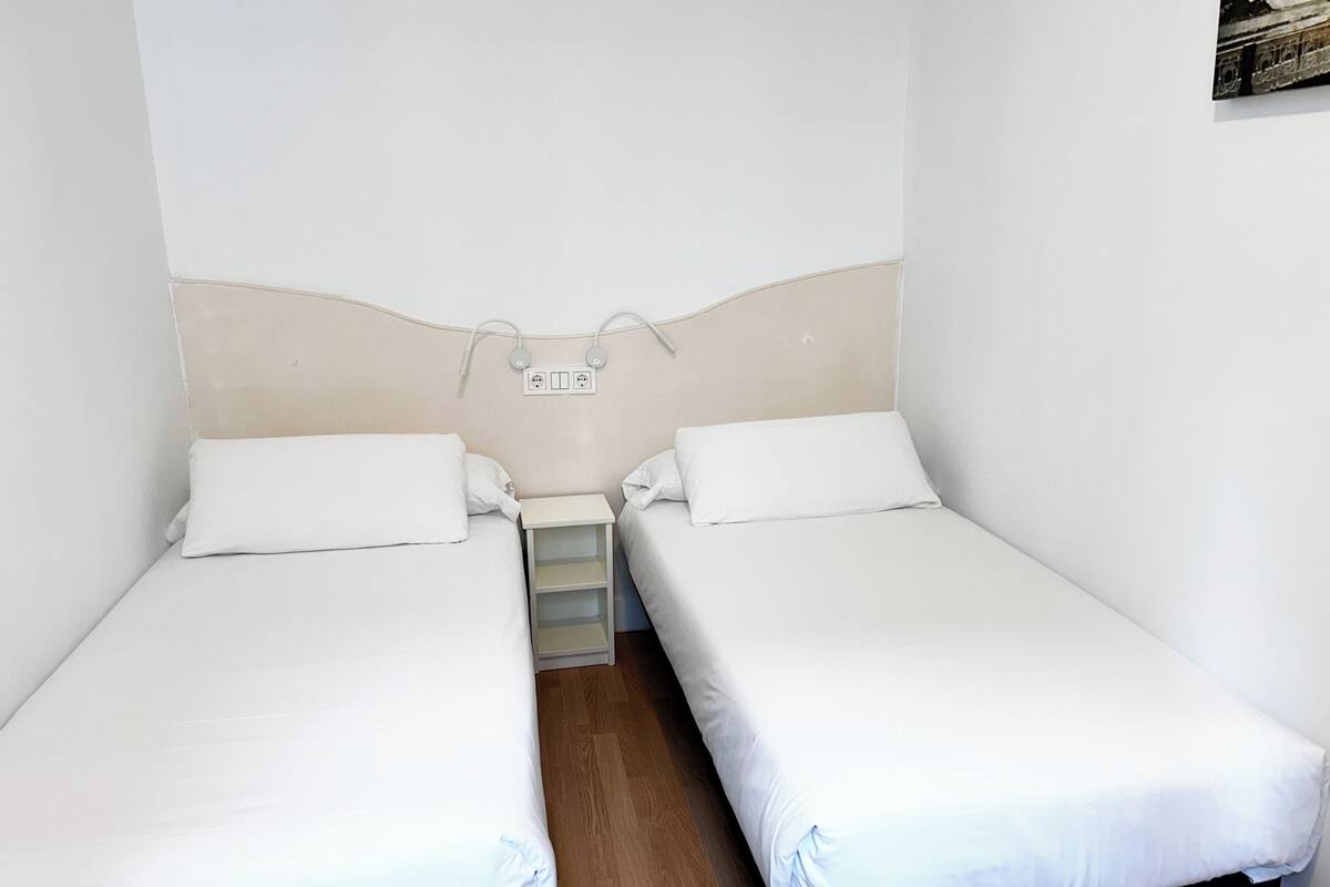 Una habitación pequeña y limpia con dos camas individuales, una mesita de noche y un enchufe en la pared.