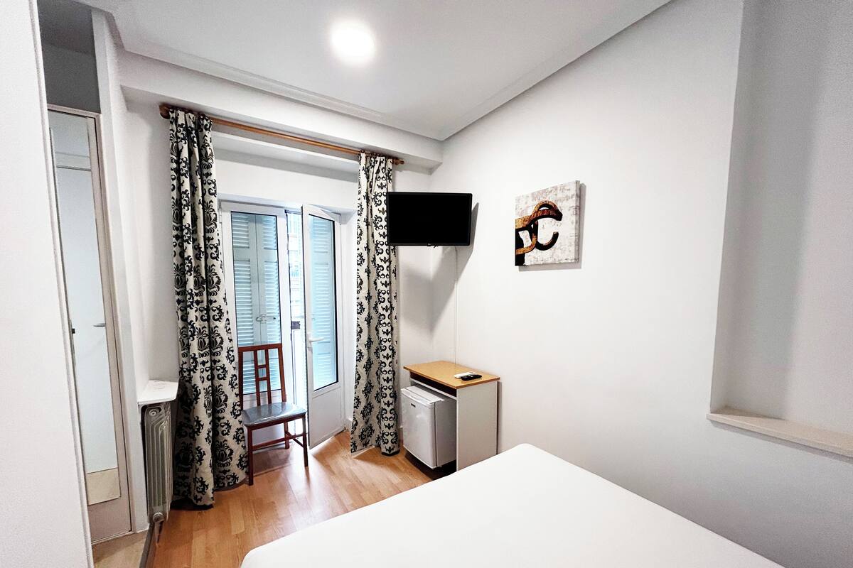 Un dormitorio con una cama, un escritorio con computadora, una silla y un televisor en la pared.