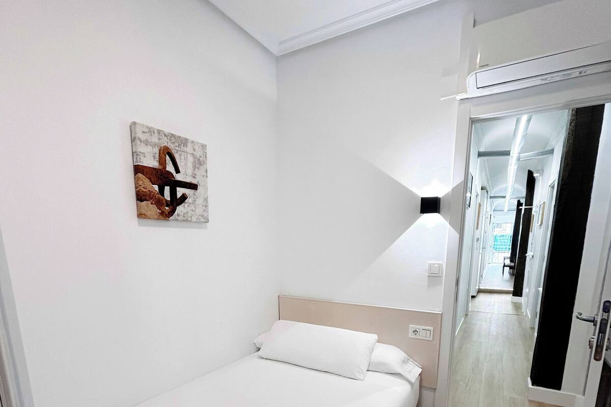 Habitación de hotel con una cama, piso de madera, pared blanca con un cuadro y una puerta que da a un pasillo.