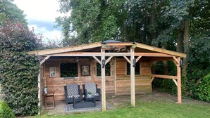 A wooden cabin with a covered patio area, two wicker chairs, and a small table with a potted plant.