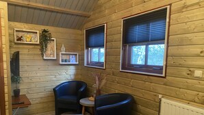 A cozy cabin interior with wooden walls, two windows with blinds, a small table with a chair, and a radiator.