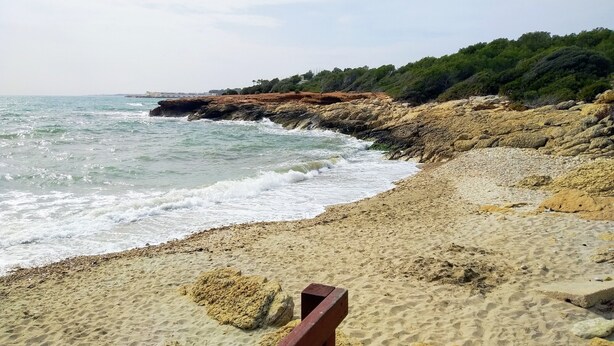 Una playa arenosa con afloramientos rocosos y una barandilla de madera.