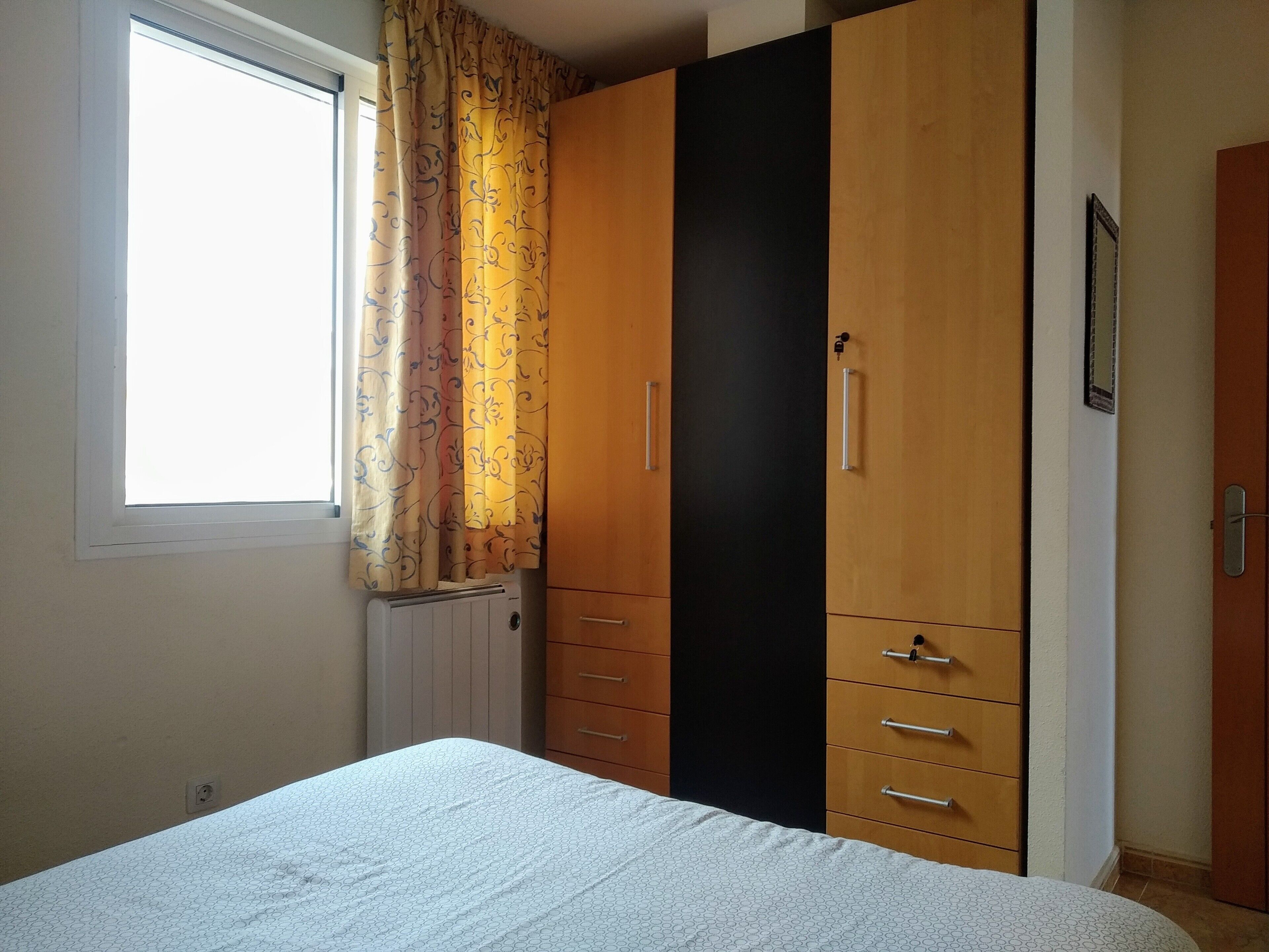 Un dormitorio con cama, armario, radiador y ventana con cortinas.