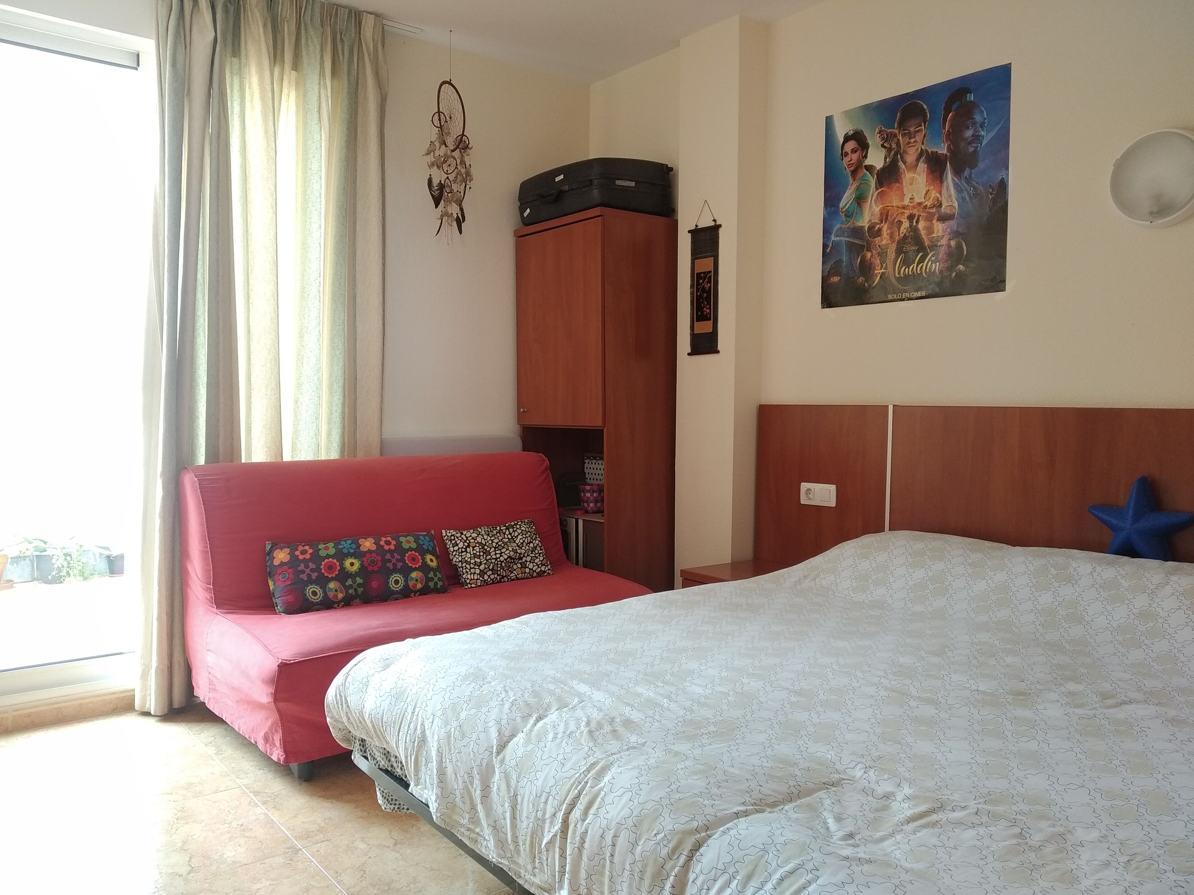 Un dormitorio con cama, sofá rojo, armario de madera, un atrapasueños y un cartel en la pared.