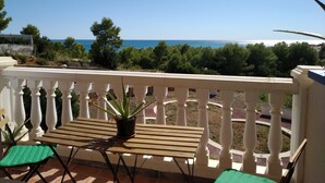 Un balcón con una mesa de madera y dos sillas, con vistas a un paisaje costero con árboles y el mar.