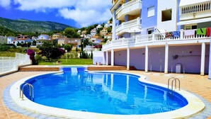 Una piscina con solárium empedrado, una zona cubierta con columnas y vistas a una colina con edificios.