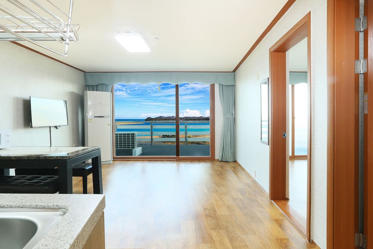 A room with a kitchen area, a dining table, a large window with a view of the sea, and a wooden door.