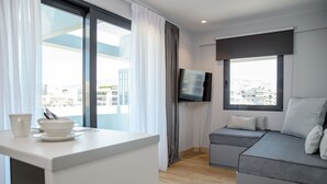 A modern hotel room with a large window, a flat-screen TV, a grey sofa, and a small table with a bowl and cups. - The Convo Syngrou Apartments (Kallithea)
