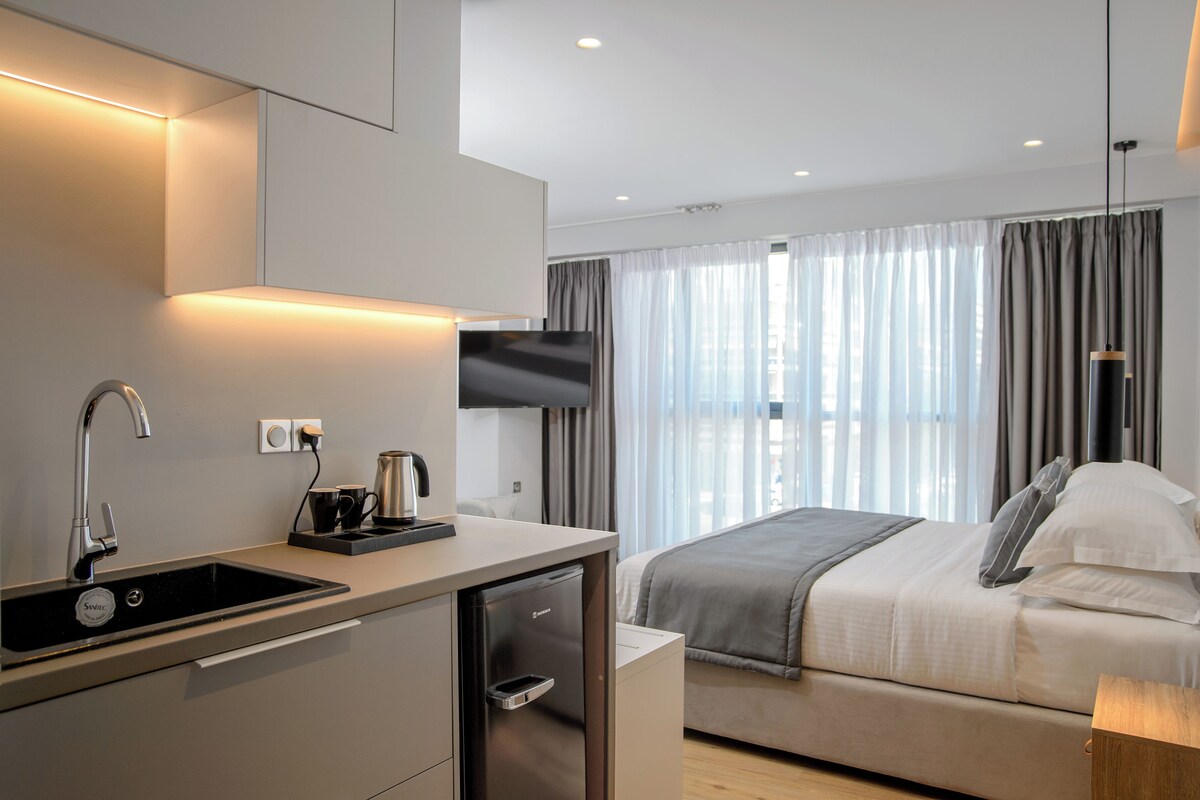 Une chambre d’hôtel moderne équipée d’une kitchenette, d’un lit avec une couverture grise et d’un téléviseur fixé au mur.