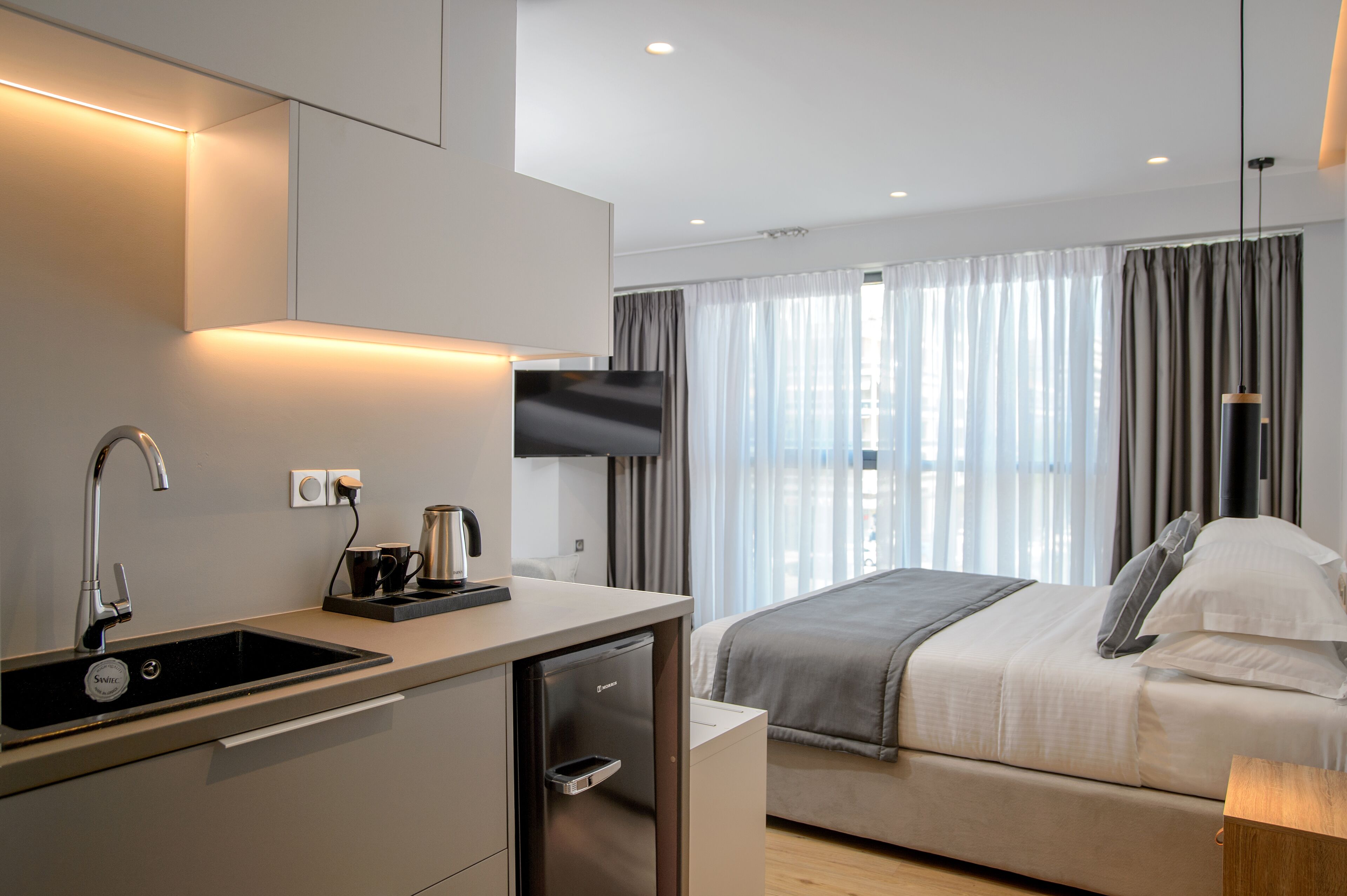 Een moderne hotelkamer met een kitchenette, een bed met een grijze deken en een aan de muur bevestigde televisie.
