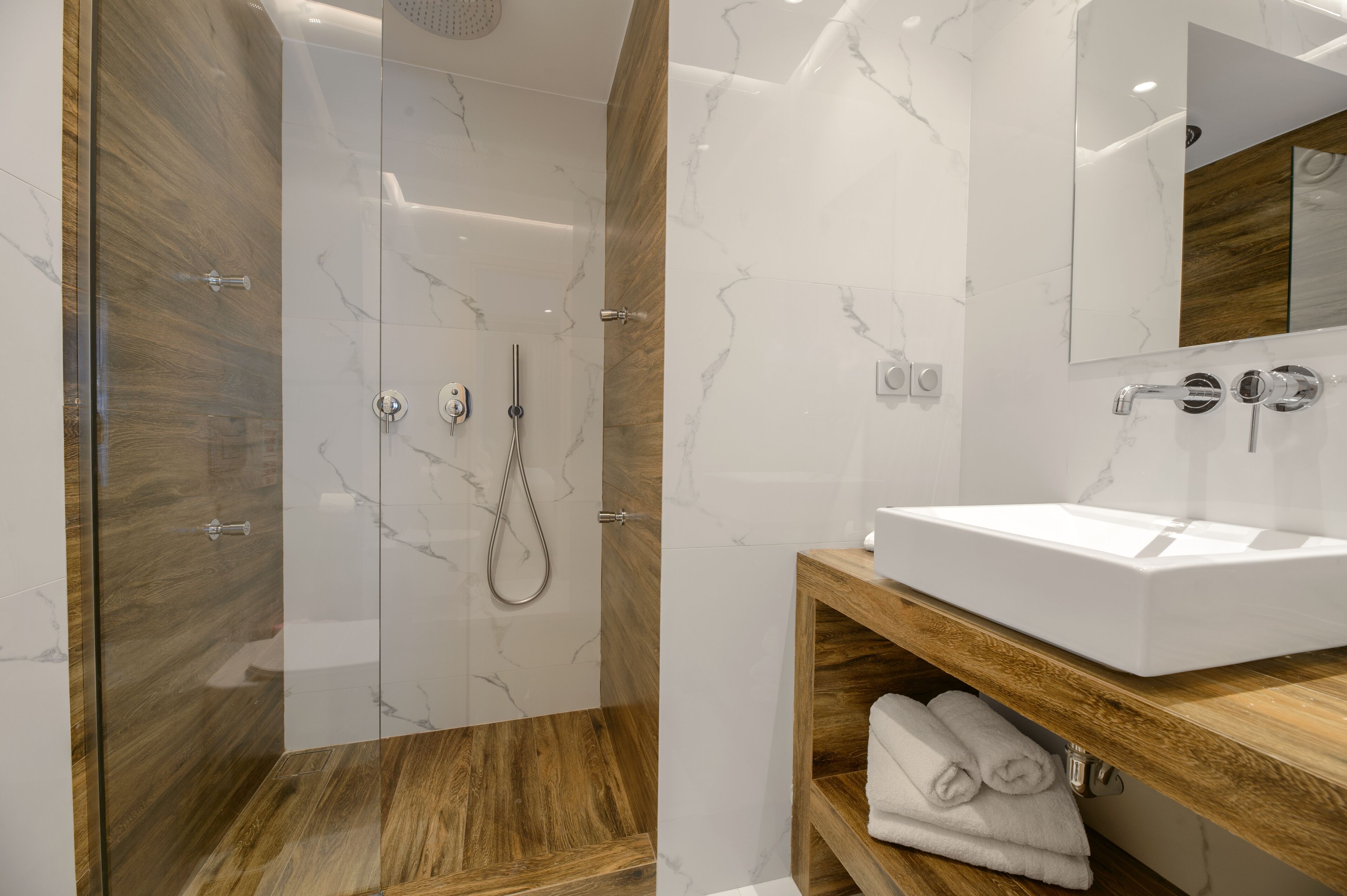 A modern bathroom with a glass-enclosed shower, a wooden vanity, and a marble-tiled wall.