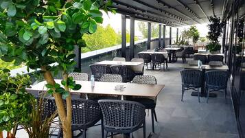 Un área de comedor al aire libre con mesas y sillas, rodeada de vegetación y un corredor cubierto.