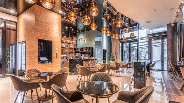 En modern hotellobby med en bardel, sittgrupper och ett stort fönster med utsikt över staden.