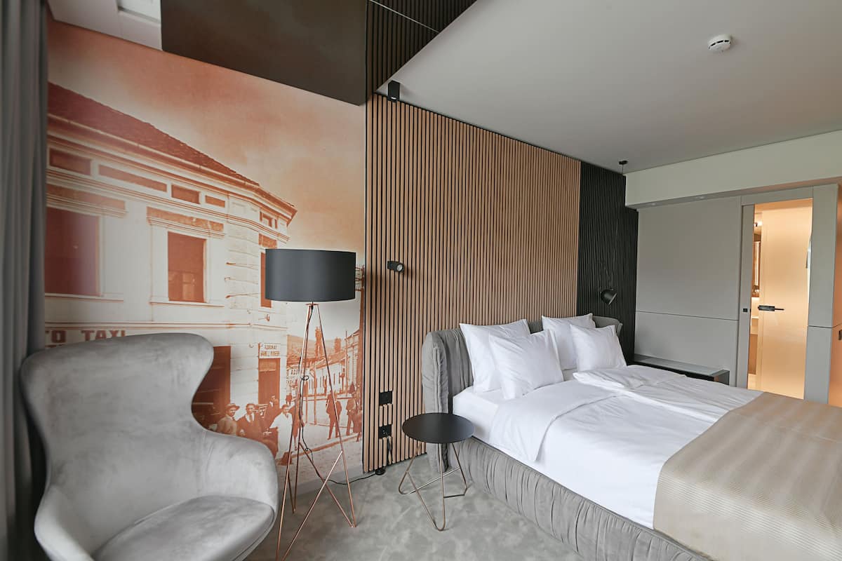 Una habitación de hotel moderna con una cama grande, un área de descanso con una silla, una mesita y una lámpara de pared.