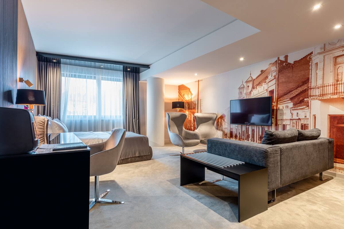 Una habitación de hotel moderna con una cama, un escritorio con una computadora portátil, un sofá gris, un televisor y un ventanal con cortinas.