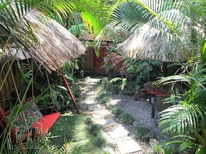A garden pathway with thatched roofs, surrounded by lush tropical plants and a small seating area.