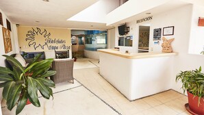 A hotel reception area with a reception desk, a sign for 'Hotel Suites Ixtapa Plaza', and a reception sign. There are potted plants and wicker furniture. - Hotel Suites Ixtapa Plaza (Ixtapa)